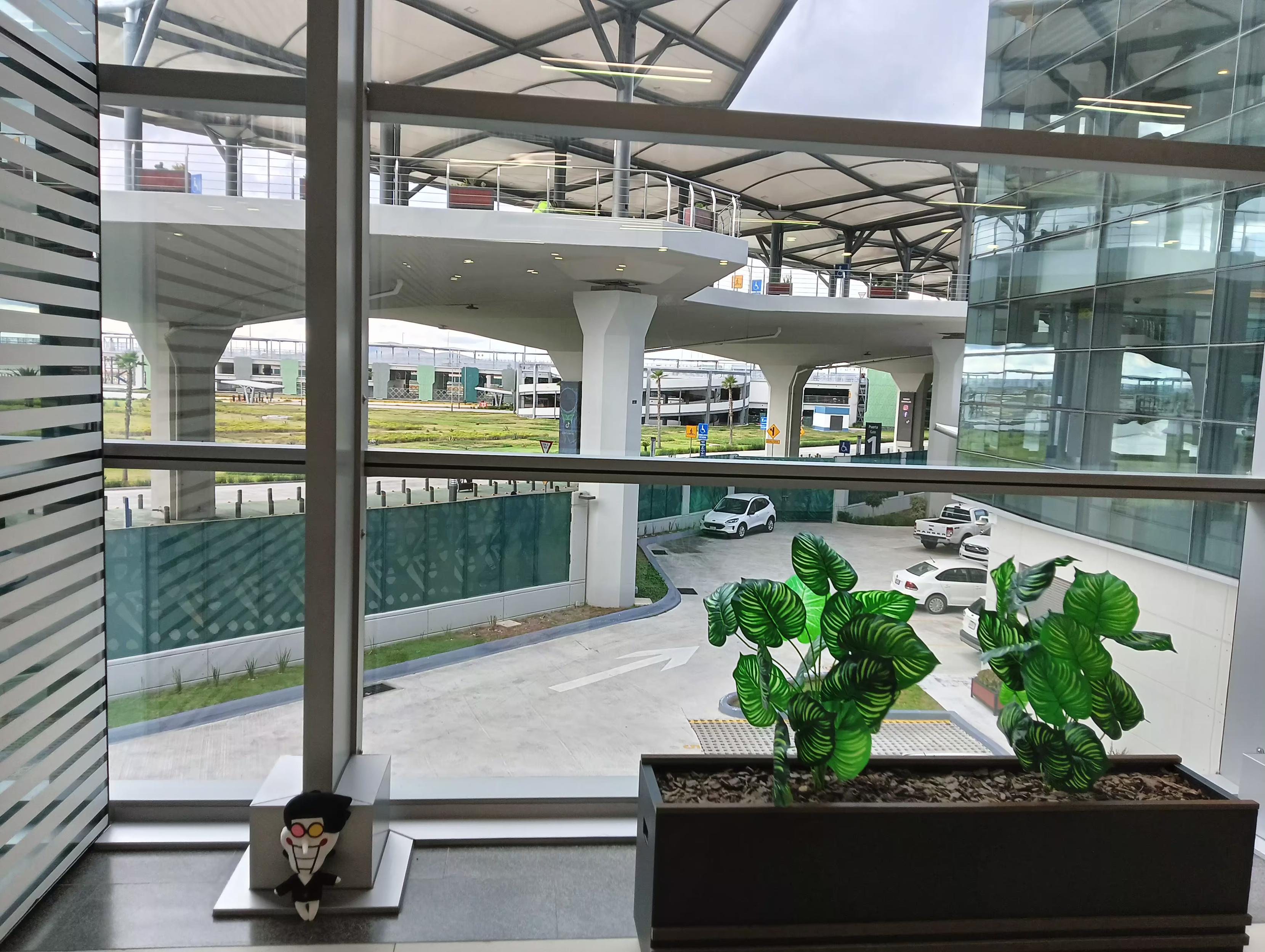 spamton en una esquina de un ventanal y junto a una maceta con plantas de plastico. la falta de gente visible le da un aura rara