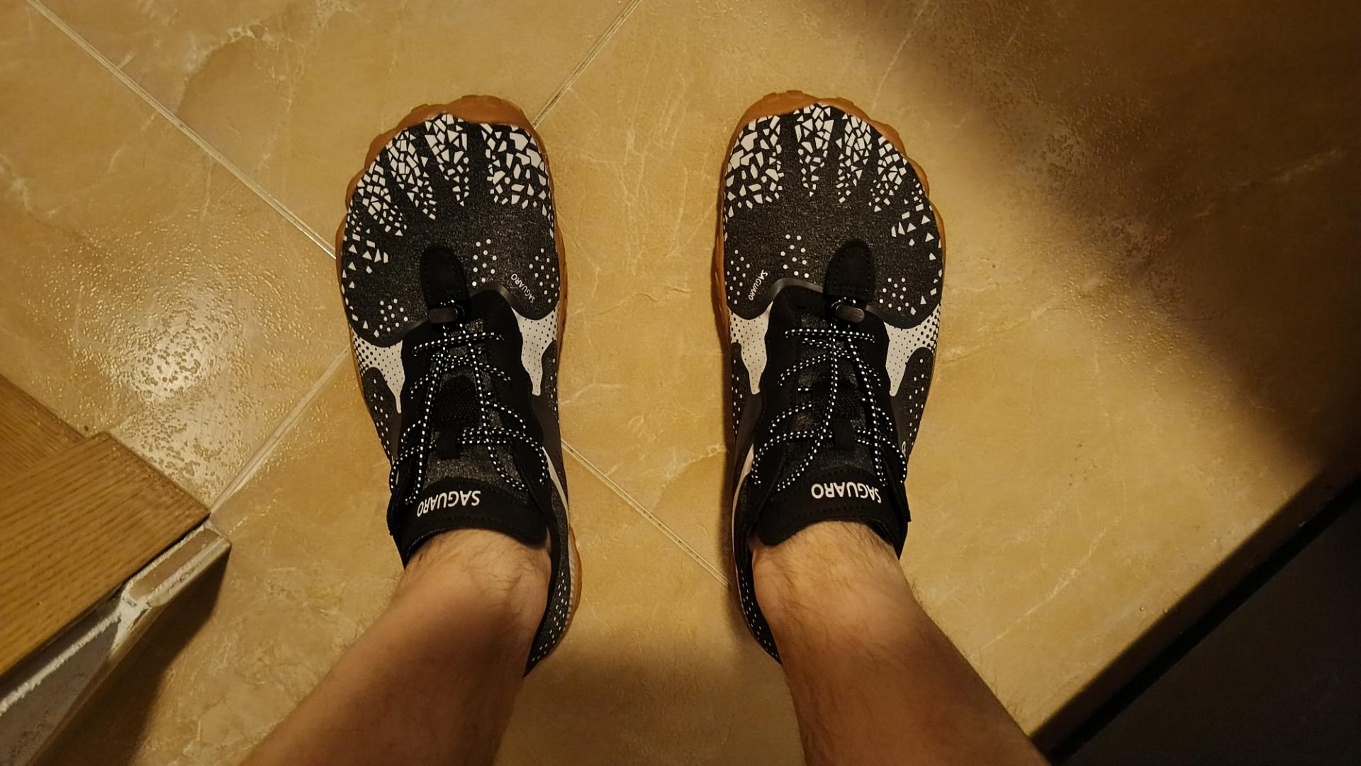 Imagen de mis pies vistos desde arriba, con las zapatillas de las que hablo. Son blancas y negras, de una tela parecida a malla elástica. La suela es de color goma, marrón claro, y tiene la zona de los dedos ligeramente ondulada, imitando su forma. La parte delantera es bastante ancha, para dejar espacio al pie.