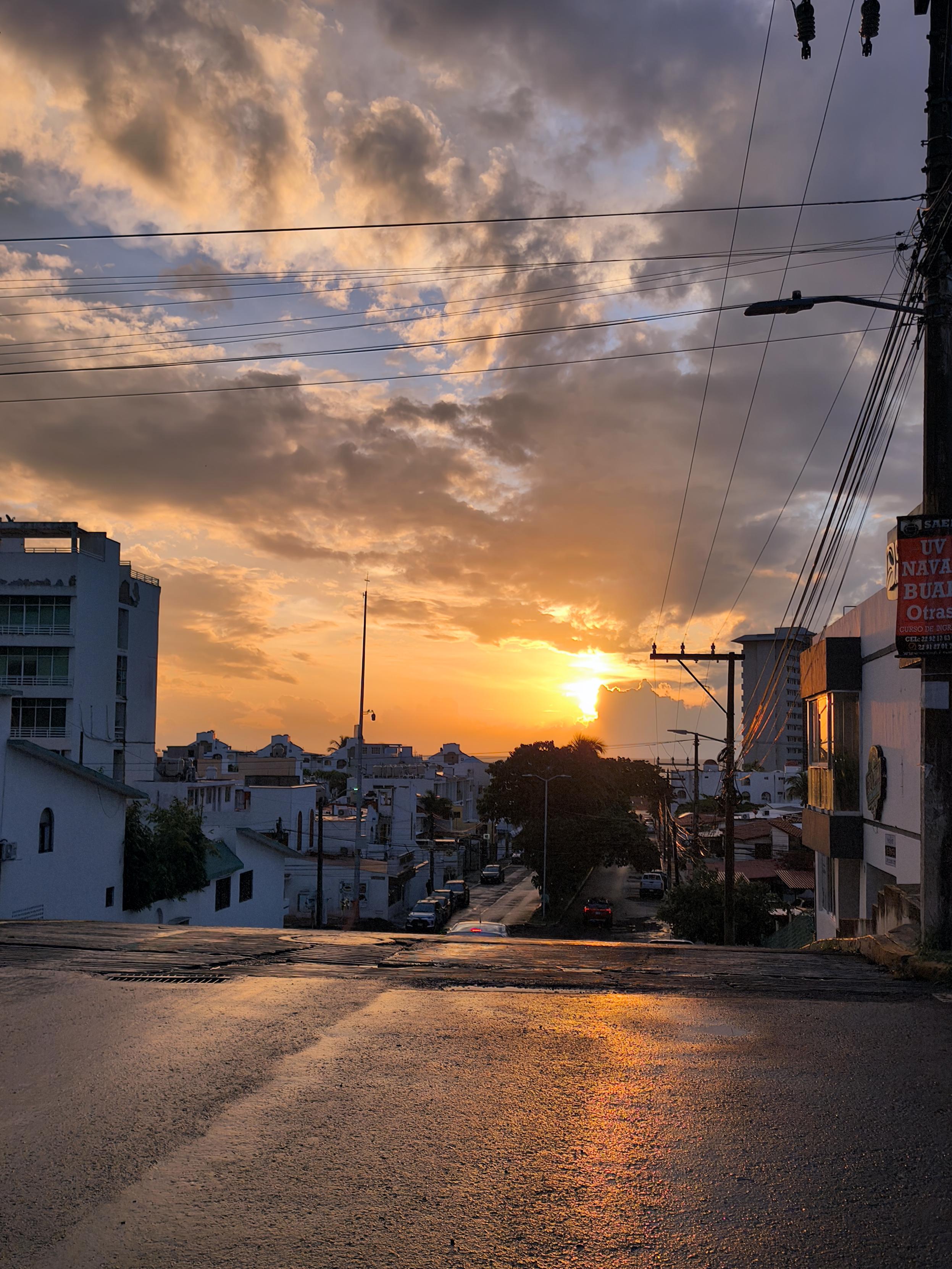 Amanecer en una calle urbana con casas y edificios a ambos lados. El sol emerge del mar al fondo, iluminando las nubes con tonos naranjas y dorados. En primer plano, la calle desciende hacia el horizonte, con vehículos estacionados, postes y cables eléctricos que atraviesan el cielo.